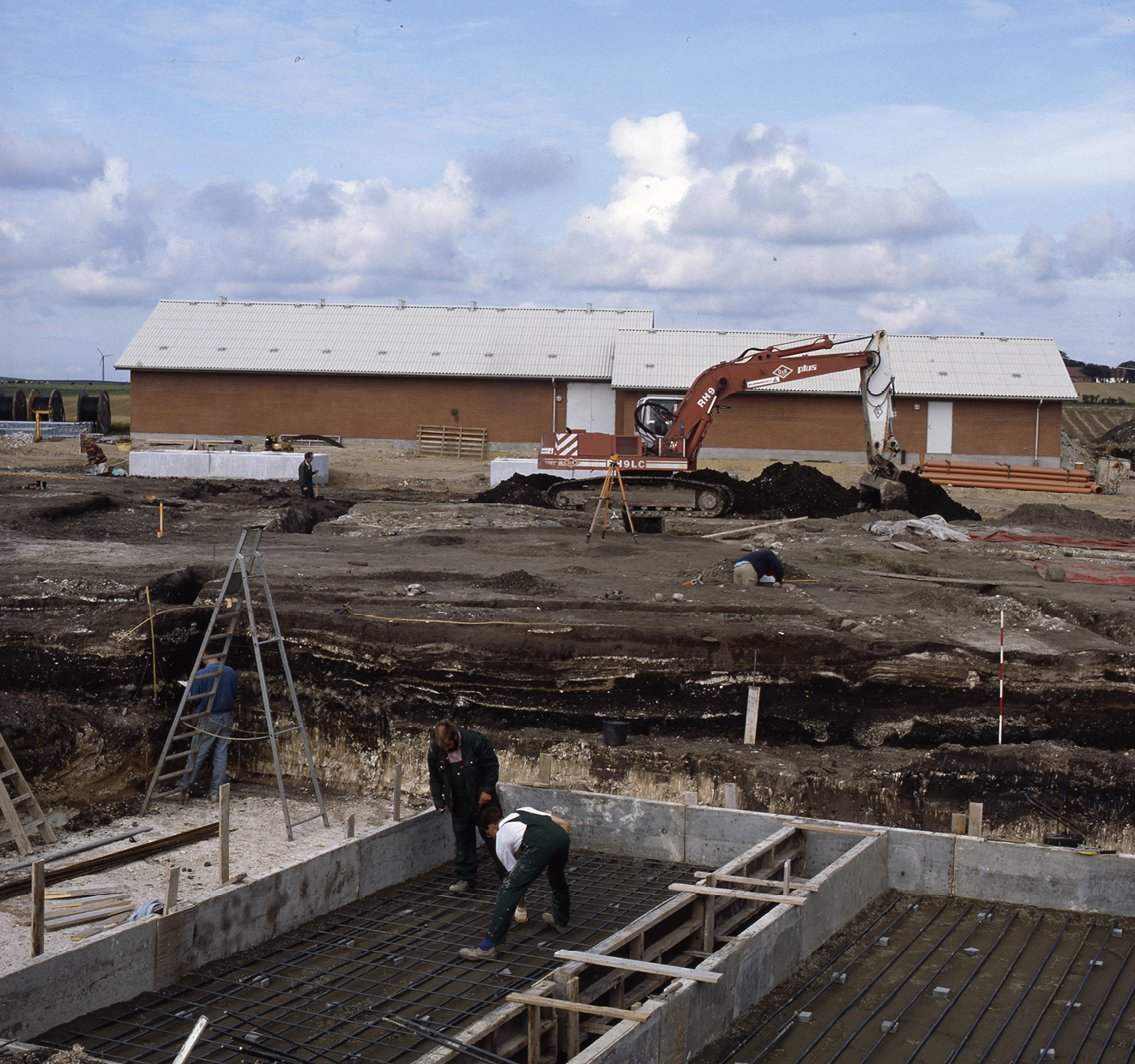 Figur 25. Foto af feltet under udgravningen set fra øst. På billedet ses Gårdkompleks C, Gård XIV og dennes forgårde og småhuse syd for forgården. I forgrunden er man allerede i gang med støbning af fundamenter til huse og i baggrund er Peter Hambro Mikkelsen i gang med profilopmålinger.